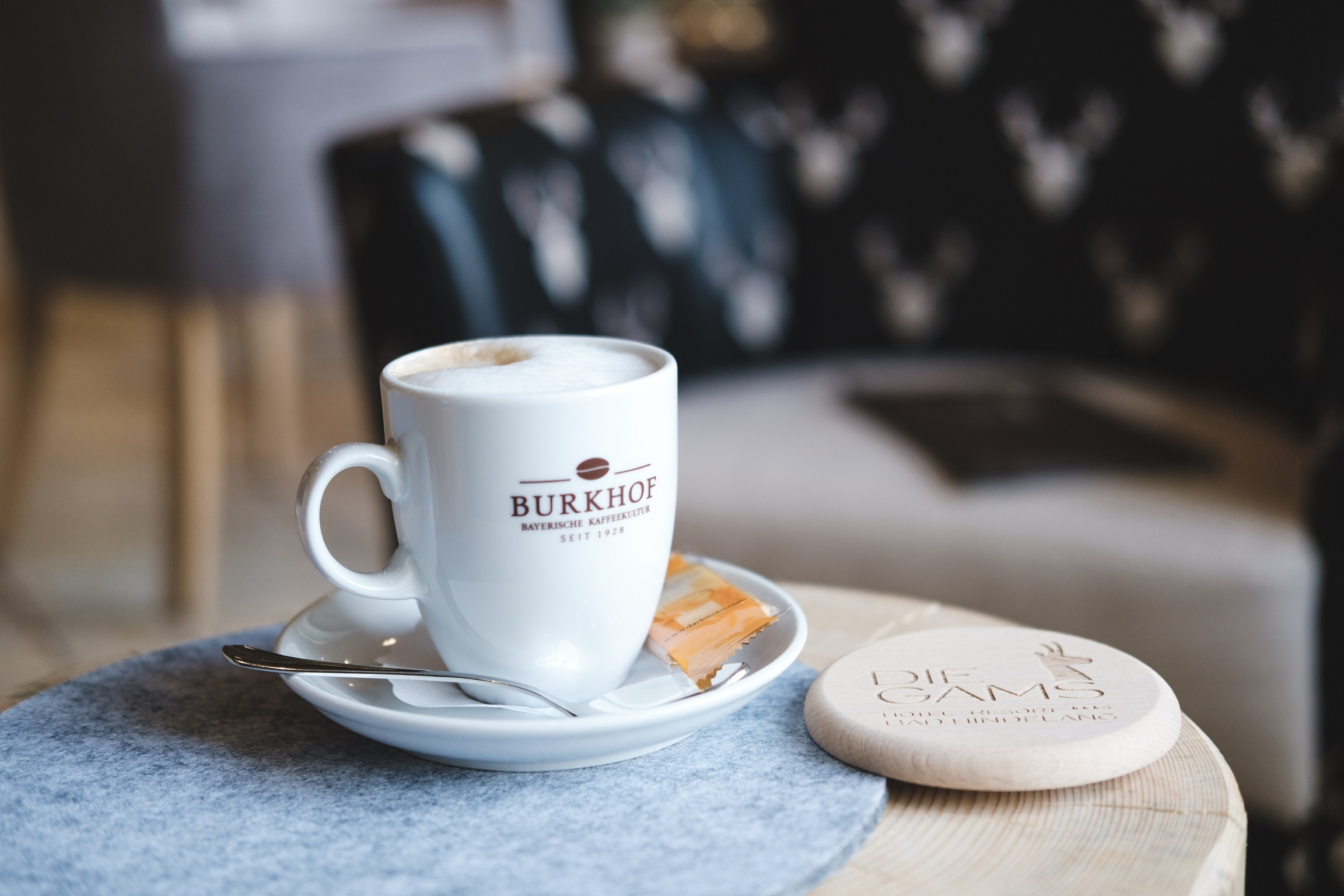 Tasse Cappuccino der Marke Burkhof mit Gebäck und Holzuntersetzer des Die Gams Hotel Resort in Bad Hindelang – gemütlicher Start in den Tag im Frühstücksbereich.