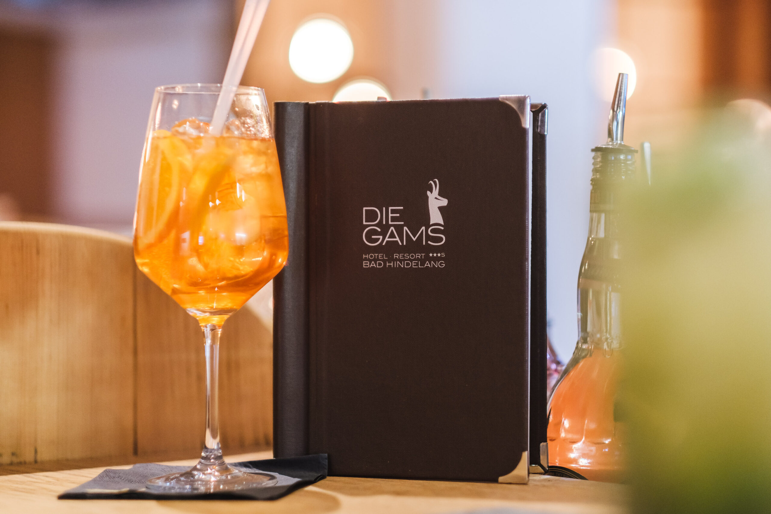Erfrischender Aperitif mit Orangenscheiben und Eis im Weinglas, serviert an der Bar des Die Gams Hotel Resort in Bad Hindelang neben stilvoller Getränkekarte.