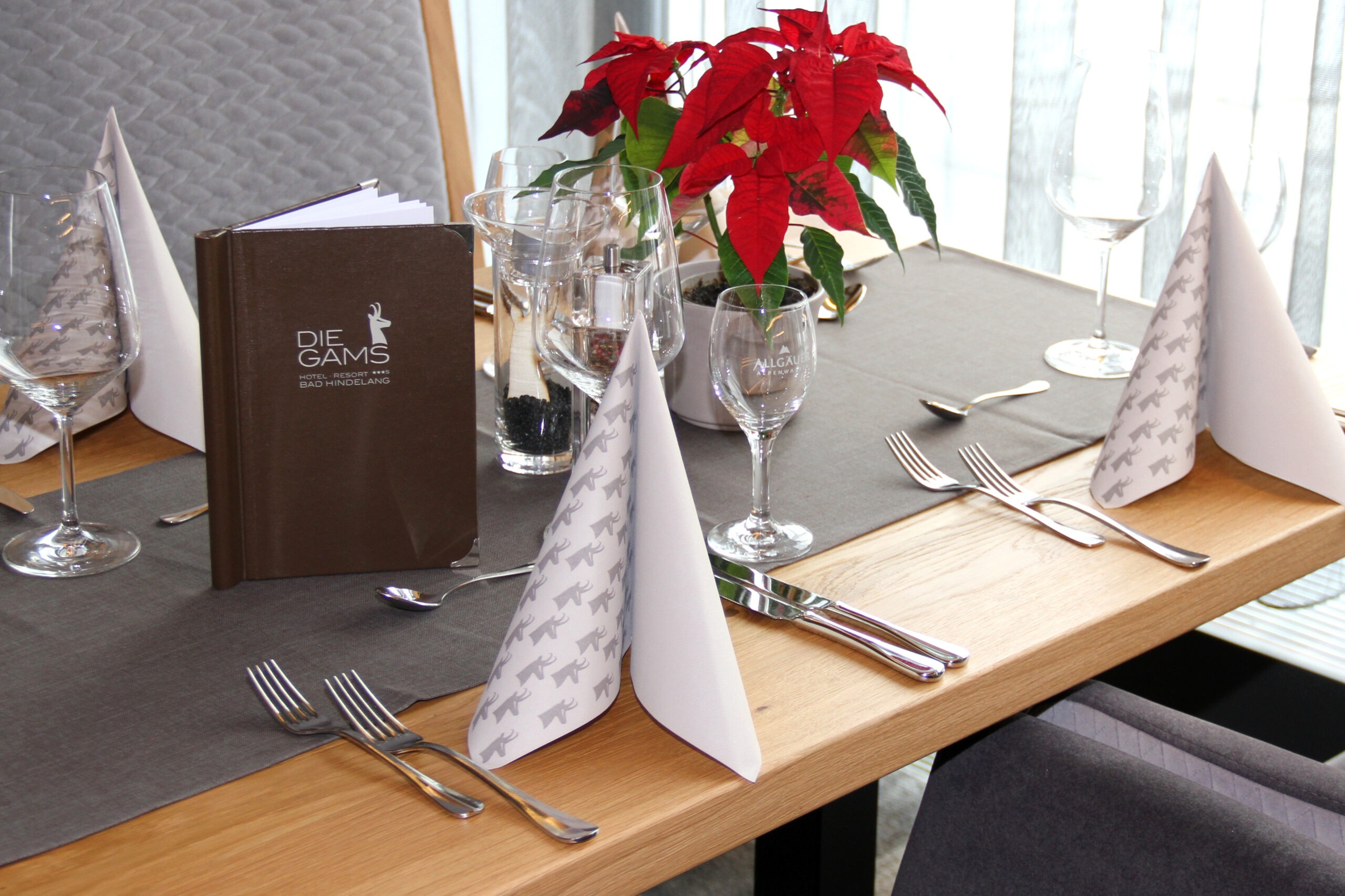 Festlich gedeckter Tisch im Die Gams Hotel Resort in Bad Hindelang mit rotem Weihnachtsstern, eleganter Menükarte und stilvoll gefalteten Servietten mit Gams-Motiv.